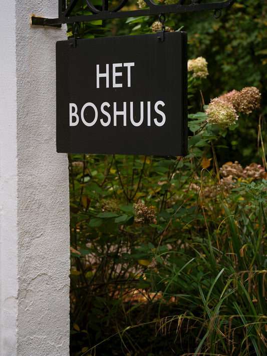 A black sign reads het boshuis with foliage behind it.